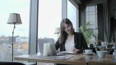 Bir not defterine notlar alan güzel bir kadın. Genç iş kadını ahşap masada kahve dükkanında oturuyor. Masada gri alüminyum dizüstü bilgisayar. Zamanlamaları ve önemli notlar yapar