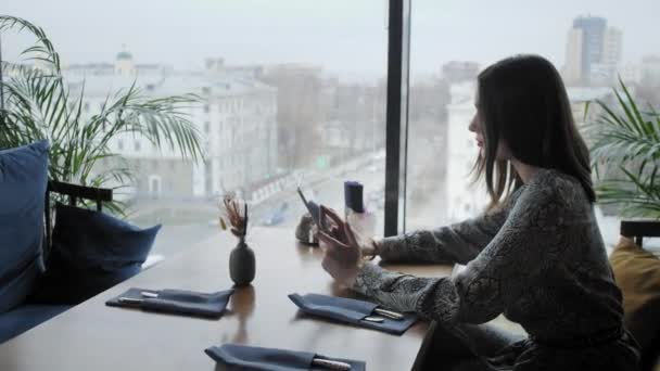 Fiatal nő gépel a táblagépen, chatel, blogol. Szabadúszó munka a kávéházban, modern coworking. Sikeres emberek, üzletember a kényelmes kávézóban. Nagy méretű ablakok — Stock videók