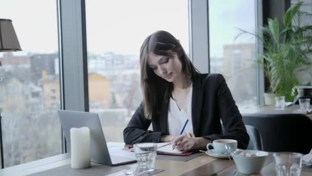 Uma mulher bonita a fazer anotações num caderno. Jovem empresária sentada em uma cafeteria à mesa de madeira. Na mesa está o laptop de alumínio cinza. Agenda e faz anotações importantes — Vídeo de Stock