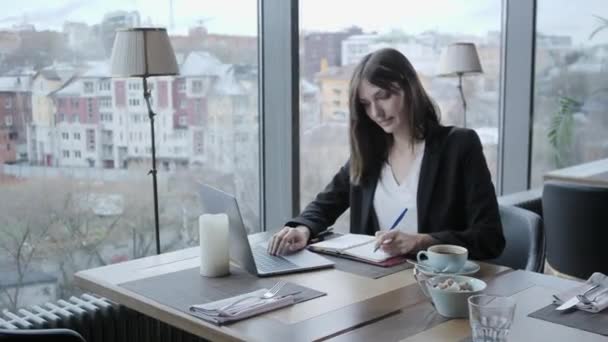 Una mujer bonita tomando notas en un cuaderno. Joven empresaria sentada en una cafetería en una mesa de madera. En la mesa hay un portátil de aluminio gris. Horarios y toma notas importantes — Vídeos de Stock