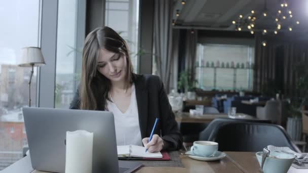 Mooie vrouw maken van notities in een notebook. Jonge zakenvrouw zittend in coffeeshop aan houten tafel. Op tafel is grijs aluminium laptop. Schema's en maakt belangrijke opmerkingen — Stockvideo