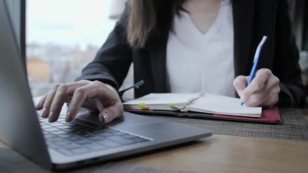 Close-up hand of a woman making notes in a notebook. Young businesswoman Sitting in coffee shop at wooden table. On table is gray aluminum laptop. Schedules and makes important notes — Stock Video