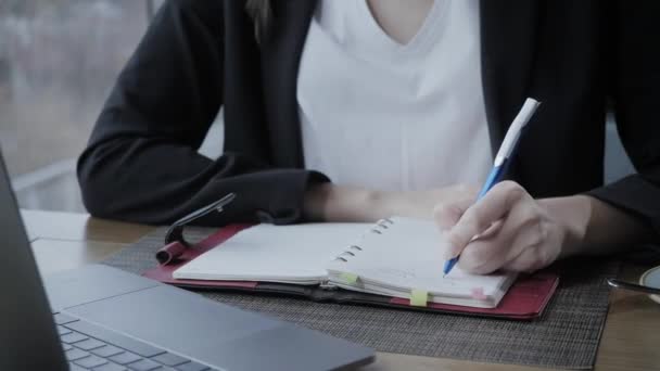 Close-up hand of a woman making notes in a notebook. Young businesswoman Sitting in coffee shop at wooden table. On table is gray aluminum laptop. Schedules and makes important notes — Stock Video