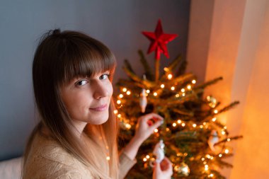 Mutlu genç kadın evde noel ağacı dekorasyon. Bir evin iç kış tatilleri. Altın ve beyaz Noel oyuncakları, ışık çelenkleri. Doğal Danimarka ladin