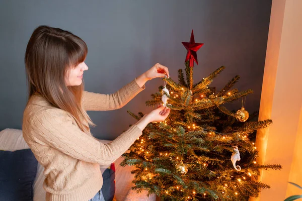 Jovem feliz decorando árvore de Natal em casa. Férias de inverno em um interior de casa. Brinquedos de Natal dourados e brancos, guirlandas de luzes. Abeto natural dinamarquês — Fotografia de Stock