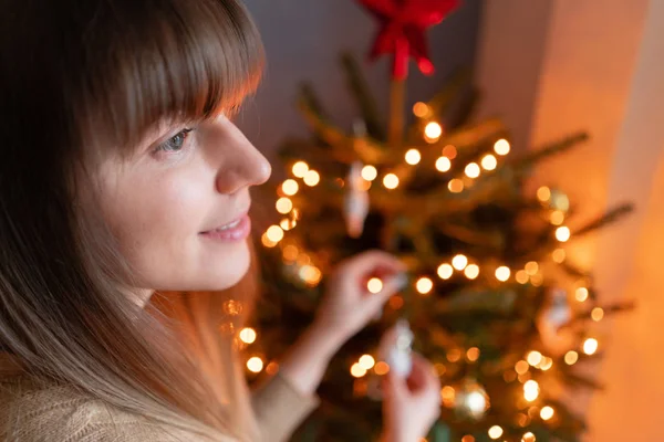 Jovem feliz decorando árvore de Natal em casa. Férias de inverno em um interior de casa. Brinquedos de Natal dourados e brancos, guirlandas de luzes. Abeto natural dinamarquês — Fotografia de Stock