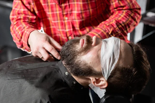 Beard cutting, face care. Barber work with clipper machine in barbershop. Professional trimmer tool cuts beard and hair of young guy in barber shop salon.
