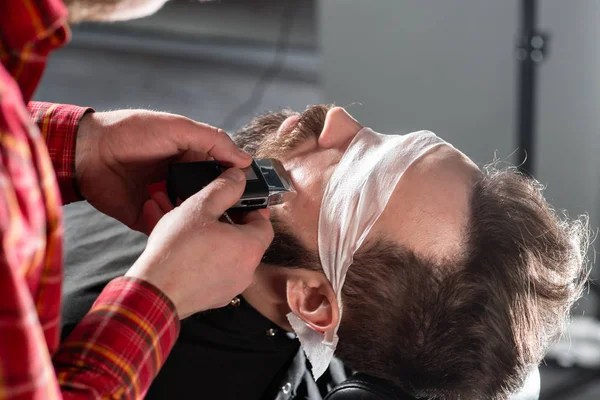 Sakal kesme, yüz bakımı. Berber dükkanında makas makinesi ile berber çalışır. Profesyonel düzeltici aracı berber dükkanı salonda genç adamın sakal ve saç keser. — Stok fotoğraf