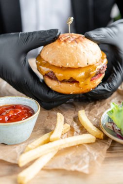 İş elbisesi ve siyah eldivenli genç adam sığır pirzolası yla lezzetli sulu çizburger yiyor. Salata ve patates kızartması, fast food ile kafede Burgers.