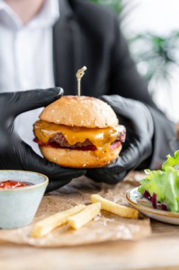 İş elbisesi ve siyah eldivenli genç adam sığır pirzolası yla lezzetli sulu çizburger yiyor. Salata ve patates kızartması, fast food ile kafede Burgers.