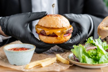 İş elbisesi ve siyah eldivenli genç adam sığır pirzolası yla lezzetli sulu çizburger yiyor. Salata ve patates kızartması, fast food ile kafede Burgers.
