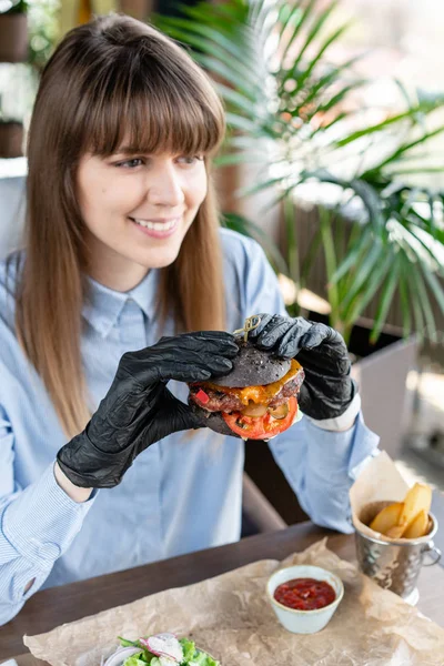 Junge Frau in blauem Hemd und schwarzen Handschuhen, die leckeren saftigen Hamburger mit Rindfleisch-Kotelett isst. Burger im Café mit Salat und Pommes, Mädchen essen Fast Food. — Stockfoto