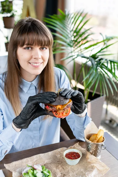 Junge Frau in blauem Hemd und schwarzen Handschuhen, die leckeren saftigen Hamburger mit Rindfleisch-Kotelett isst. Burger im Café mit Salat und Pommes, Mädchen essen Fast Food. — Stockfoto