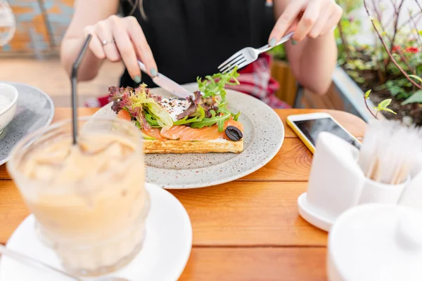 Traditional Belgian waffle with salmon, lettuce leaves and poached egg. A young woman is having Breakfast in a summer cafe, hands with a fork and a knife close-up. Cold coffee with ice cubes — Stock Photo, Image