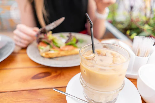 Focus in foreground on Cold coffee with ice cubes. A young woman is having Breakfast in a summer cafe. — Stock Photo, Image