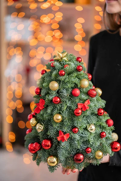 Piccolo bellissimo albero di Natale Realizzato a mano. Fiorista di lavoro. Disposizione di abete fresco, oro e palline rosse. L'umore natalizio. Bokeh di ghirlanda luci su sfondo . — Foto Stock