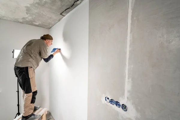 Молодой ремонтник выравнивается с пластиковой лопаткой. Рабочий приклеивает обои к бетонной стене. Ремонт квартиры. Концепция ремонта дома. Белые обои для краски — стоковое фото