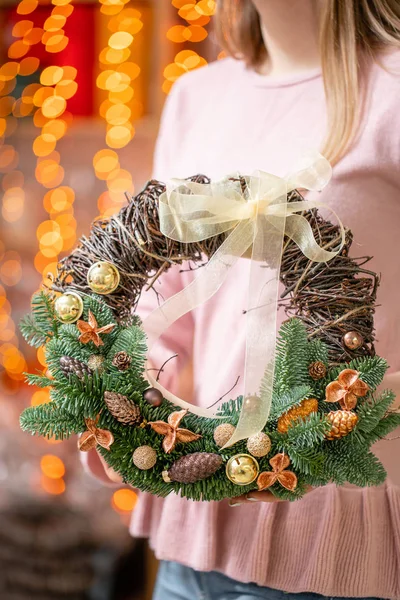 Bella corona festiva di abete rosso fresco con giocattoli a sfera nelle mani della donna. L'umore natalizio. Bokeh di ghirlanda luci su sfondo . — Foto Stock