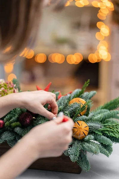 Mujer joven crea y decora hermoso arreglo festivo de abeto fresco, ornamentales en una caja de madera rústica. Humor de Navidad. Guirnalda bokeh en el fondo . — Foto de Stock