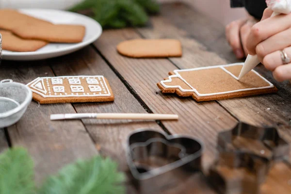 Helado de panadería navideña. Mujer decorando galletas de miel de jengibre sobre mesa marrón de madera. Primer plano, espacio de copia. Galleta en blanco casa de jengibre, listo para decorar . — Foto de Stock