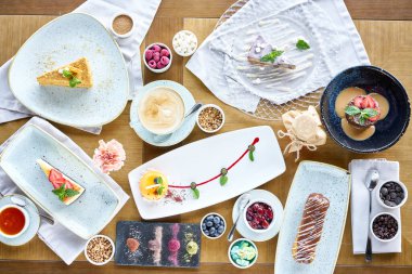 Kafede kahvaltı, sabah kahvesi. Kapuçino ve masada bir sürü tatlı var. Ekmek, kremalı pasta, çikolatalı mantar ve peynirli kek.,