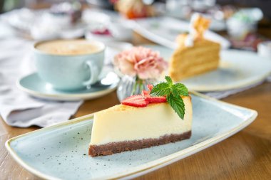 Beyaz tabakta çilekli ve nane yapraklı lezzetli bir cheesecake. Kafede kahvaltı, sabah kahvesi. Cappuccino ve masada bir sürü tatlı..