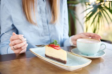 Genç bir kadın bir restoranda kahve kapuçino içer ve tatlı yer. Beyaz tabakta çilekli ve nane yapraklı lezzetli bir cheesecake. Restoran menüsü