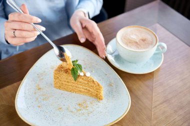Genç bir kadın bir restoranda kahve kapuçino içer ve tatlı yer. Krema ve nane yapraklarıyla süslenmiş bal süngeri pastası. Kafede kahvaltı, sabah kahvesi. Restoran menüsü