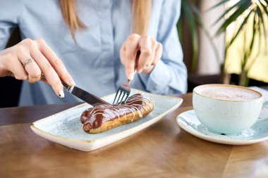Genç bir kadın bir restoranda kahve kapuçino içer ve tatlı yer. Geleneksel Fransız ekleride çikolata. Vanilya kremalı ve çikolatalı pasta..
