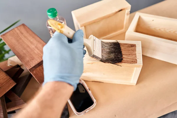 Hombre carpintero barnizado caja de madera para flores con pincel en su taller de carpintería pequeña empresa. En su trabajo, ¿utiliza manchas o conservantes de madera para mostrar el patrón de madera . — Foto de Stock