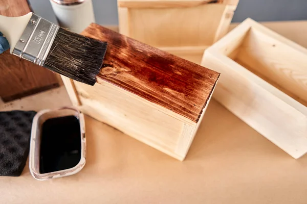 Hombre carpintero barnizado caja de madera para flores con pincel en su taller de carpintería pequeña empresa. En su trabajo, ¿utiliza manchas o conservantes de madera para mostrar el patrón de madera . — Foto de Stock