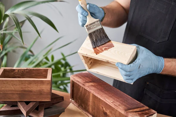 Чоловік-тесляр лакує дерев'яну ящик для квітів пензлем у своїй майстерні з деревообробки малого бізнесу. У своїй роботі, ви використовуєте плями або консерванти для деревини, щоб показати візерунок деревини . — стокове фото