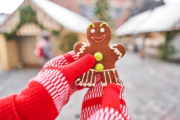 Hand in red mitten holding a smiling gingerbread man and christmas mood in blurred background. Christmas market in old town European small city.