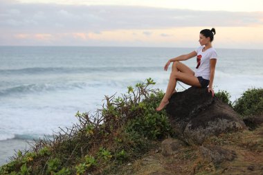 Yana doğru taş uçurumdan deniz ve gökyüzü arka planı üzerinde oturan poz kız
