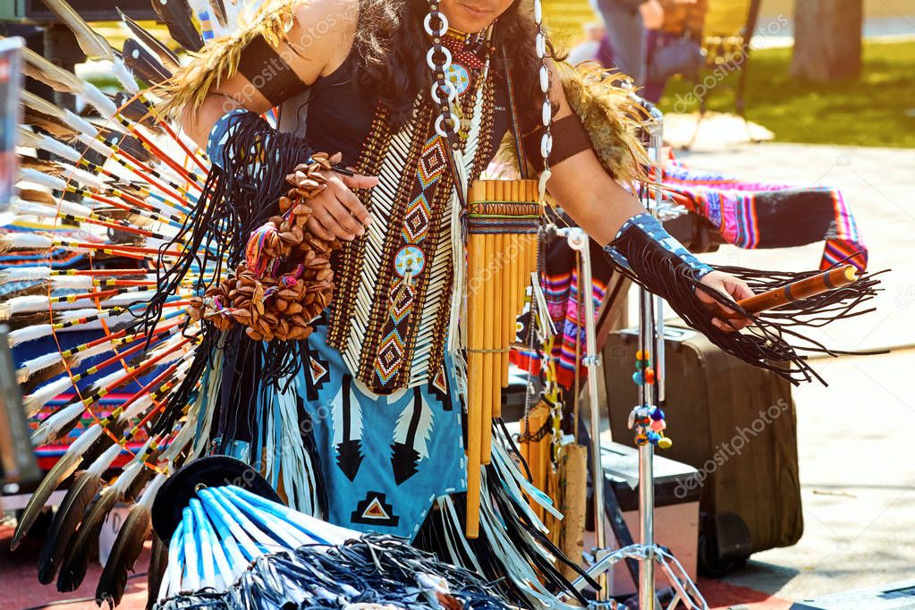 An Indian from South America dances in a national costume with feathers, with a flute in the street in the summer on a clear day.