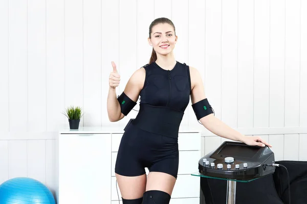 A beautiful young girl stands next to the simulator for practicing electro stimulation of muscles EMS training. The concept of modern fitness using high technologies.