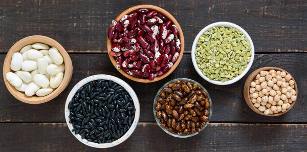 Healthy food, dieting, nutrition concept, vegan protein source. Assortment of colorful raw legumes: green peas, beans, chickpeas in bowls, on a wooden table. Top view. Banner.