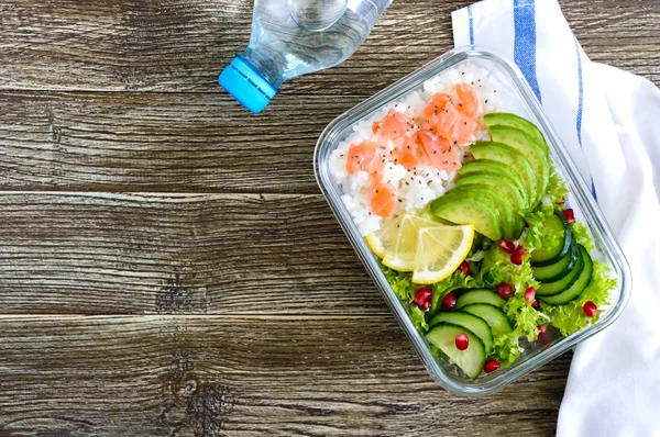 Lunch box: rice, salmon, salad with cucumber, avocado, greens, lemon, chia seeds and bottle of water on a wooden background. Fitness food. The concept of healthy eating. School lunch box. Top view, flat lay. Copy space.