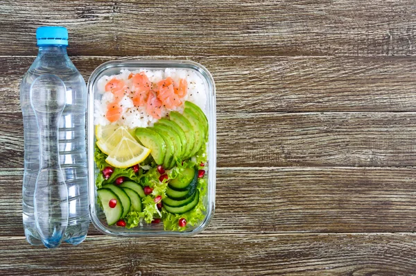 Lunch box: rice, salmon, salad with cucumber, avocado, greens, lemon, chia seeds and bottle of water on a wooden background. Fitness food. The concept of healthy eating. School lunch box. Top view, flat lay. Copy space.