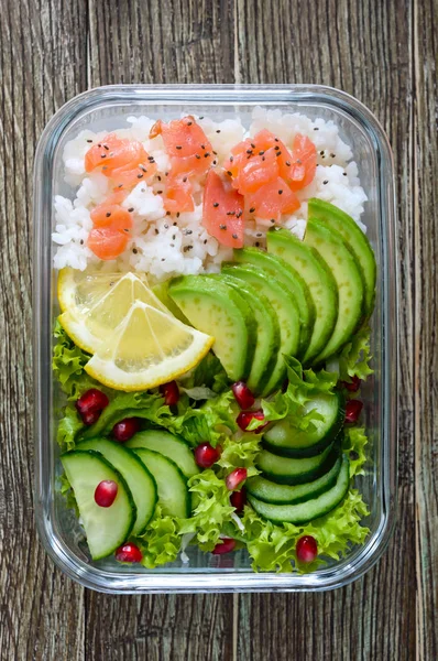 Lunch box: rice, salmon, salad with cucumber, avocado, greens, lemon, chia seeds on a wooden background. Fitness food. The concept of healthy eating. School lunch box. Top view, flat lay. Close up.