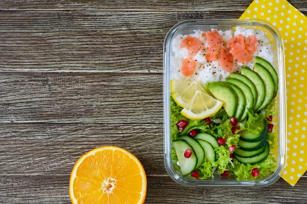 Lunch box: rice, salmon, salad with cucumber, avocado, greens, lemon, fresh orange, and bottle of water on a wooden background. Fitness food. The concept of healthy eating. School lunch box. Copy space