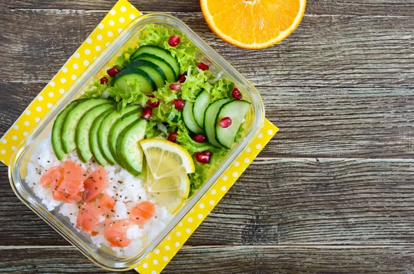 Lunch box: rice, salmon, salad with cucumber, avocado, greens, lemon, fresh orange, and bottle of water on a wooden background. Fitness food. The concept of healthy eating. School lunch box. Copy space