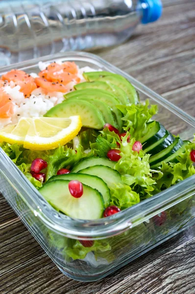 Lunch box: rice, salmon, salad with cucumber, avocado, greens, lemon, fresh orange, and bottle of water on a wooden background. Fitness food. The concept of healthy eating. School lunch box