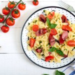 Smakelijke pasta Farfalle met gegrilde worstjes, verse cherry tomaten en basilicum op een bord op een witte houten achtergrond. Bovenaanzicht, vlakke lay.