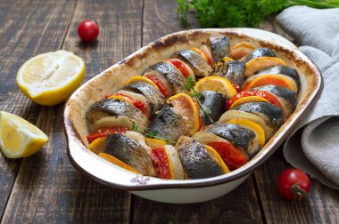 Baked fish with vegetables. Pieces of herring, onions, tomato, lemon, aromatic herbs in ceramic form on a rustic wooden table clipart