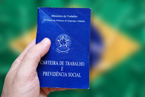 Man's hand holding Brazilian Work Card isolated on brazilian flag defocused background. Written in Portuguese Federative Republic of Brazil, Ministry of Labor. Work and Social Security card. Brazilian work concept