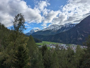 Karlı dağlarla kaplı bir dağ manzarası, yeşil bir vadi, küçük bir köy, sık ormanlar ve parçalı bulutlu bir gökyüzünün altındaki yüksek ağaçlar..
