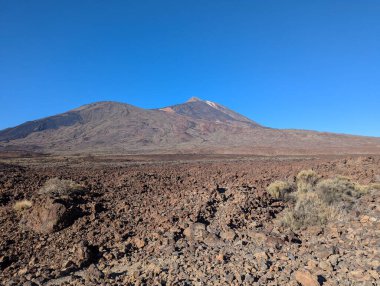 Kanarya Adaları 'ndaki Tenerife' nin volkanik zirvesi olan Teide Dağı 'nın geniş bir manzarası var. Önplan boyunca uzanan engebeli lav alanları var. Görüntü, açık ve aydınlık bir günde, Teide Ulusal Parkı 'nın keskin volkanik arazisini ve doğal güzelliğini yakalıyor..