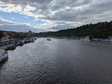 Çek Cumhuriyeti 'nin Prag kentindeki Vltava Nehri manzaralı bir manzarada nehir kenarına yanaşmış tekneler, ağaçlarla kaplı tepeler ve dramatik bulutların altındaki tarihi gökyüzü. Bu görüntü Prag 'ın nehir kenarındaki cazibesini yansıtıyor.
