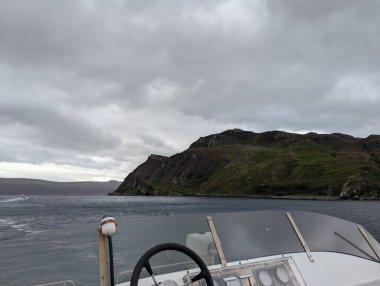 Bir tekne dümeni, bulutlu bir gökyüzü altında engebeli yeşil tepelere ve Raasay 'ın Sesi' nin kayalıklarına doğru manzarayı çerçeveler. Karanlık sakin su, uzak kara kütleleri yoğun bulutların arasından çıkarken karamsar atmosferi yansıtır..